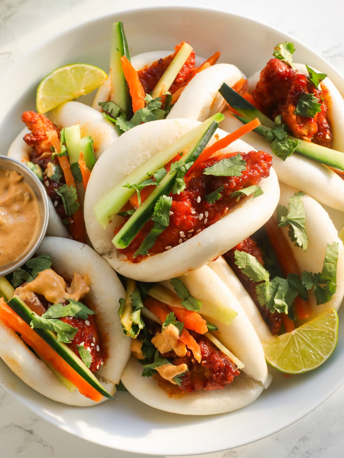 Korean chicken served with bao buns, cucumber, carrots on a white plate with sauce.