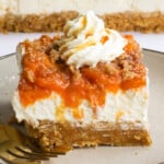 Peaches cheesecake bar with whipped cream, streusel and caramel on top of a brown plate with a gold fork.