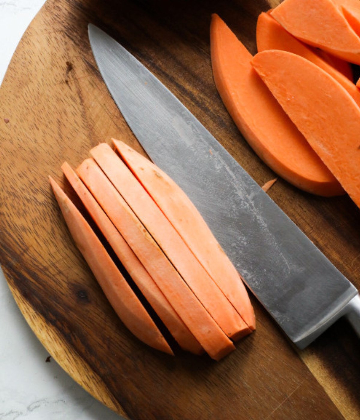 Sliced sweet potatoes on a wooden chopping board and a silver chefs knife.