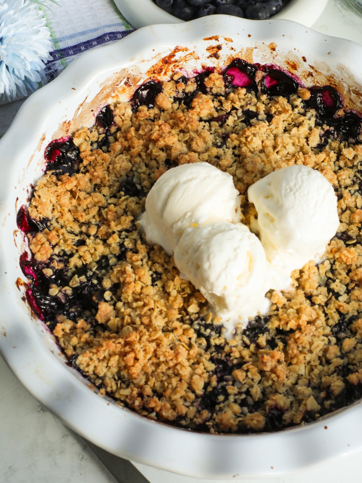 Blueberry crisp in a white pie dish with ice cream served on top.