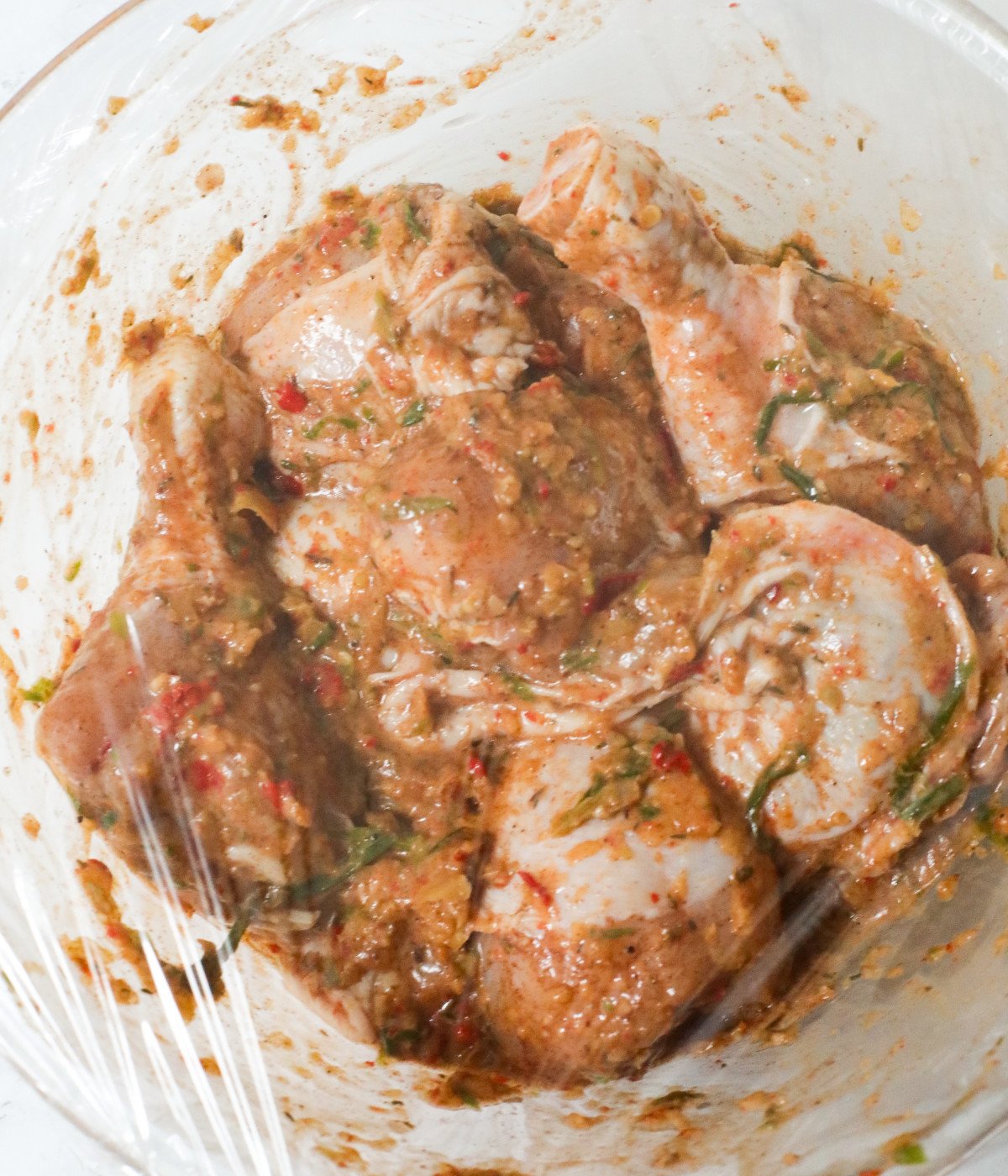 Uncooked thighs, drums and jerk marinade in a glass bowl covered with plastic wrap.