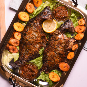 Two whole grilled tilapia in a tray with plantain, lettuce and pepper sauce.