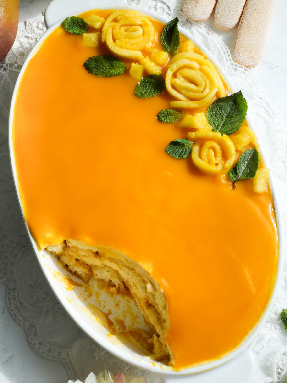 Mangomisu in a oval white dish with mango pulp, mango roses and mint leaves on top.