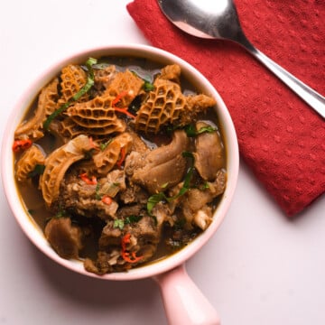 Tripe, goat meat pepper soup in a pink bowl garnished with chillies and spinach. A spoon and red cloth.