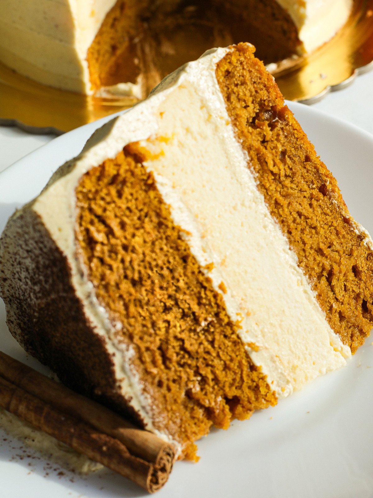A slice of pumpkin cheese cake cake with frosting on a white plate with a cinnamon stick.