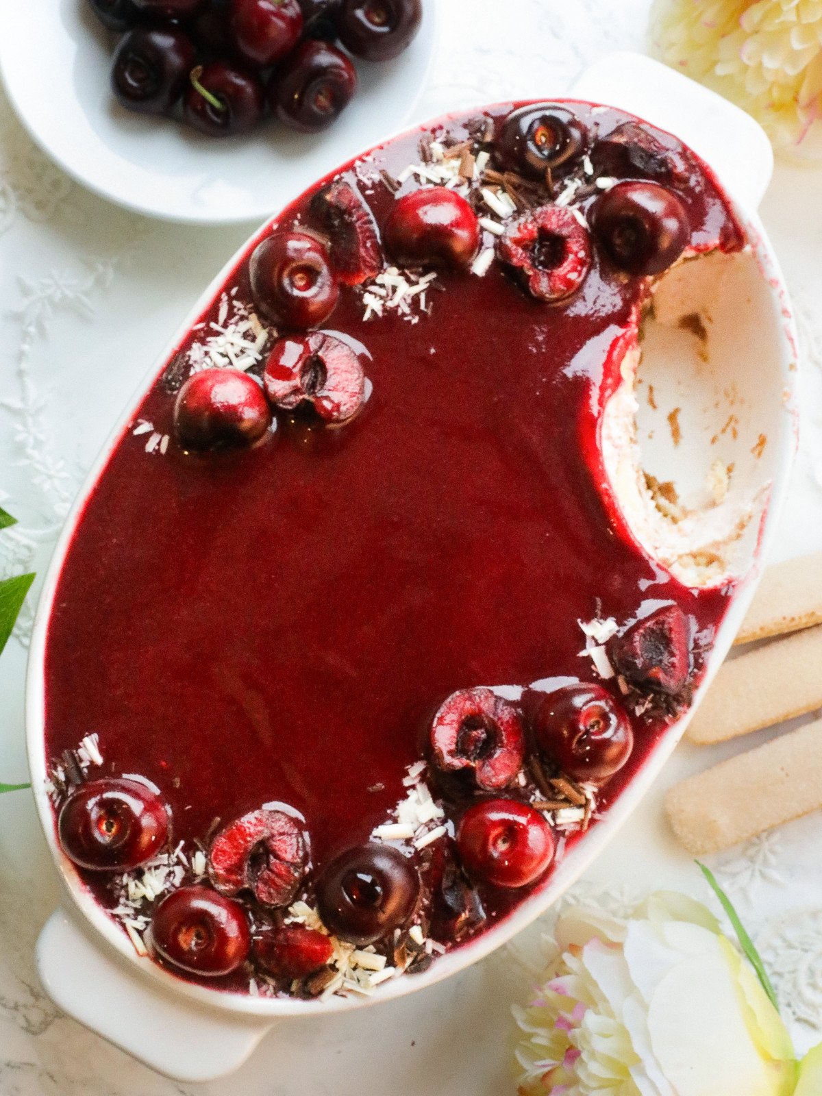 Cherries tiramisu in a white oval dish on a white table with a slice missing.