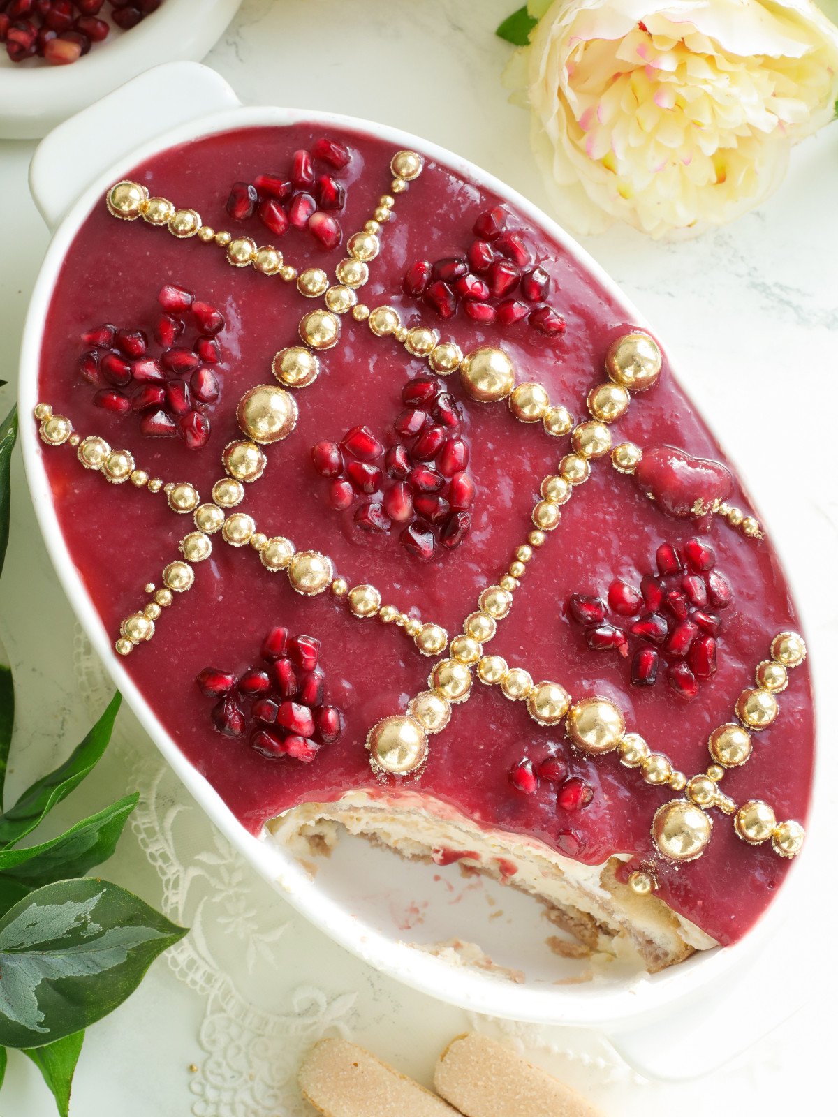 Pomegranate tiramisu with diagonal gold sprinkles and heart shaped pomegrante arils in a white oval dish.