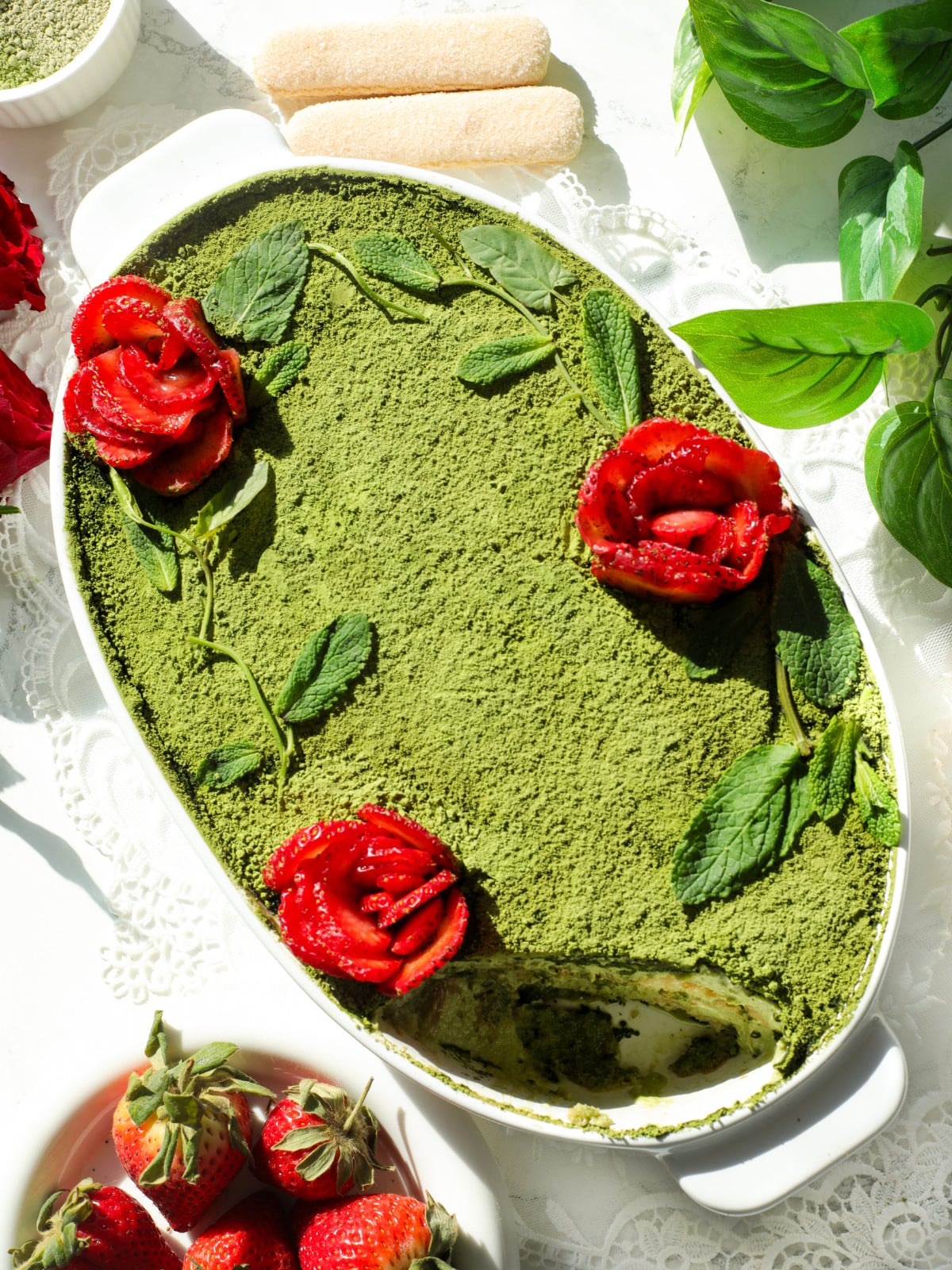 Matcha with strawberry tiramisu in a white oval dish with roses and matcha powder on top.